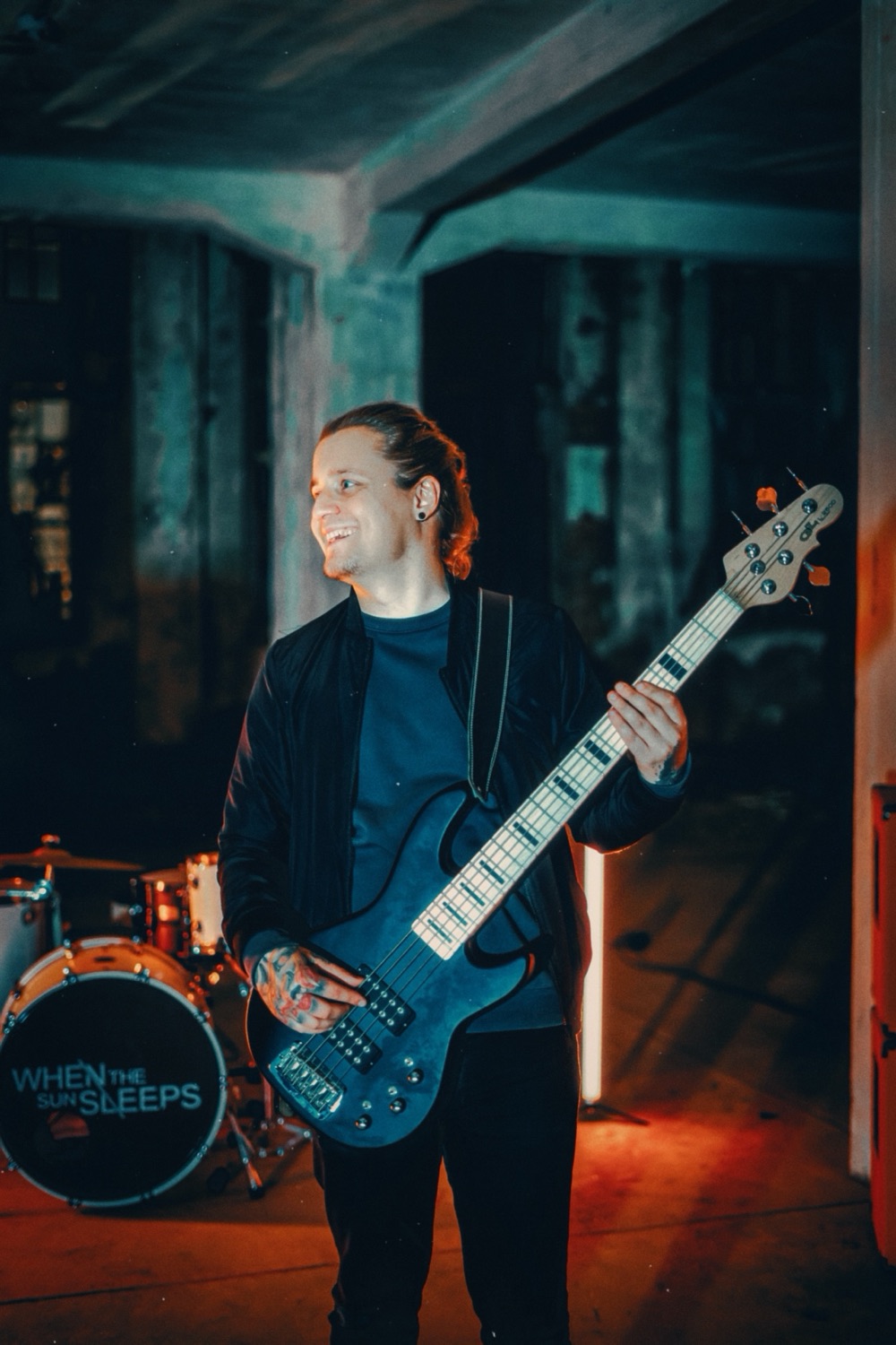 Sascha Herrmann lacht und hält einen fünfsaitigen E-Bass, im Hintergrund ein Schlagzeug.